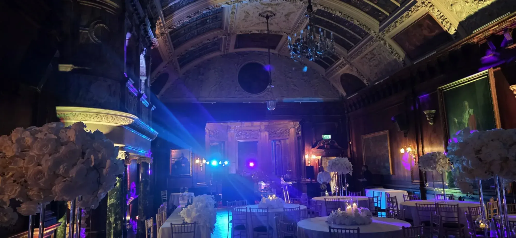 Blue and purple event lighting in an ornate reception room with floral table arrangements