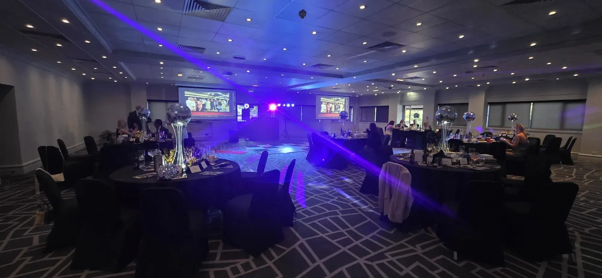 A function room arranged with round tables, projection screens and blue DJ lighting