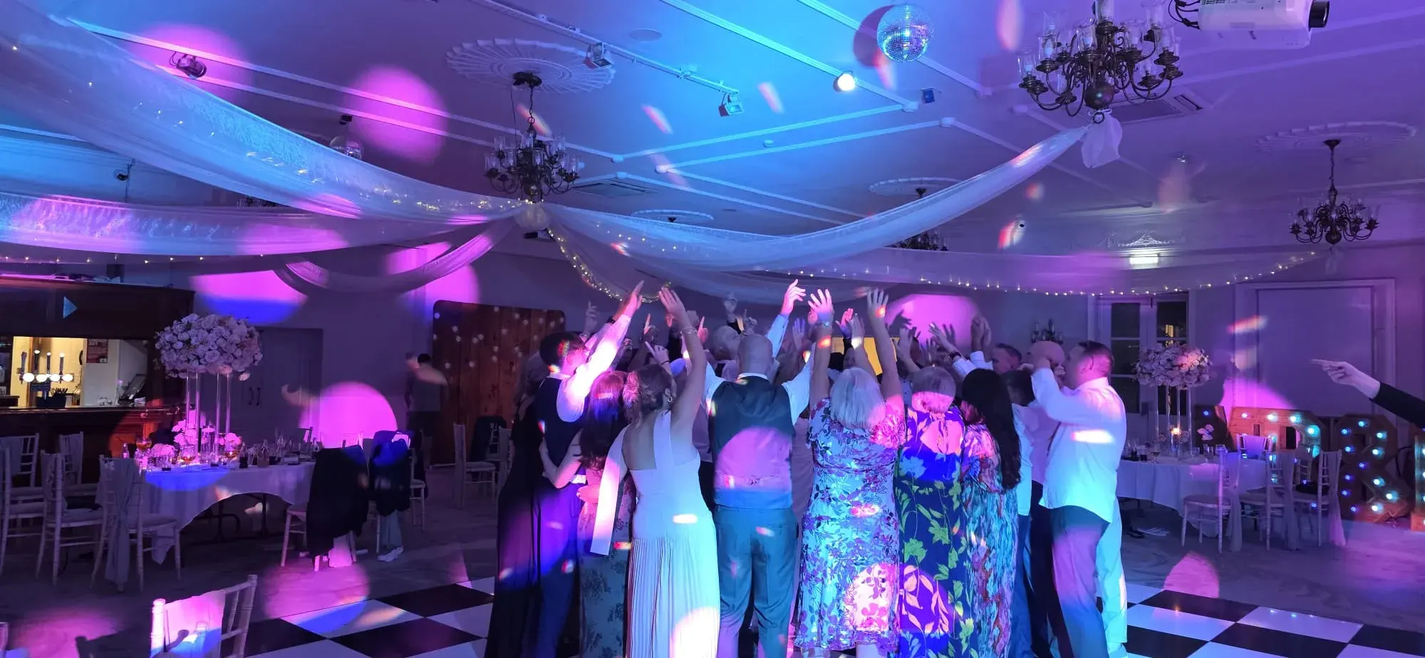 A crowd of wedding guests raising their hands beneath blue and pink party lighting