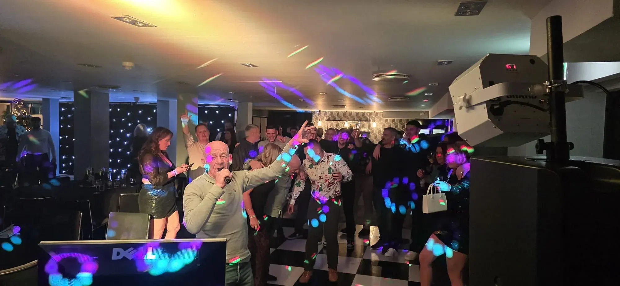 A party guest singing into a microphone with one hand raised and a crowd behind him