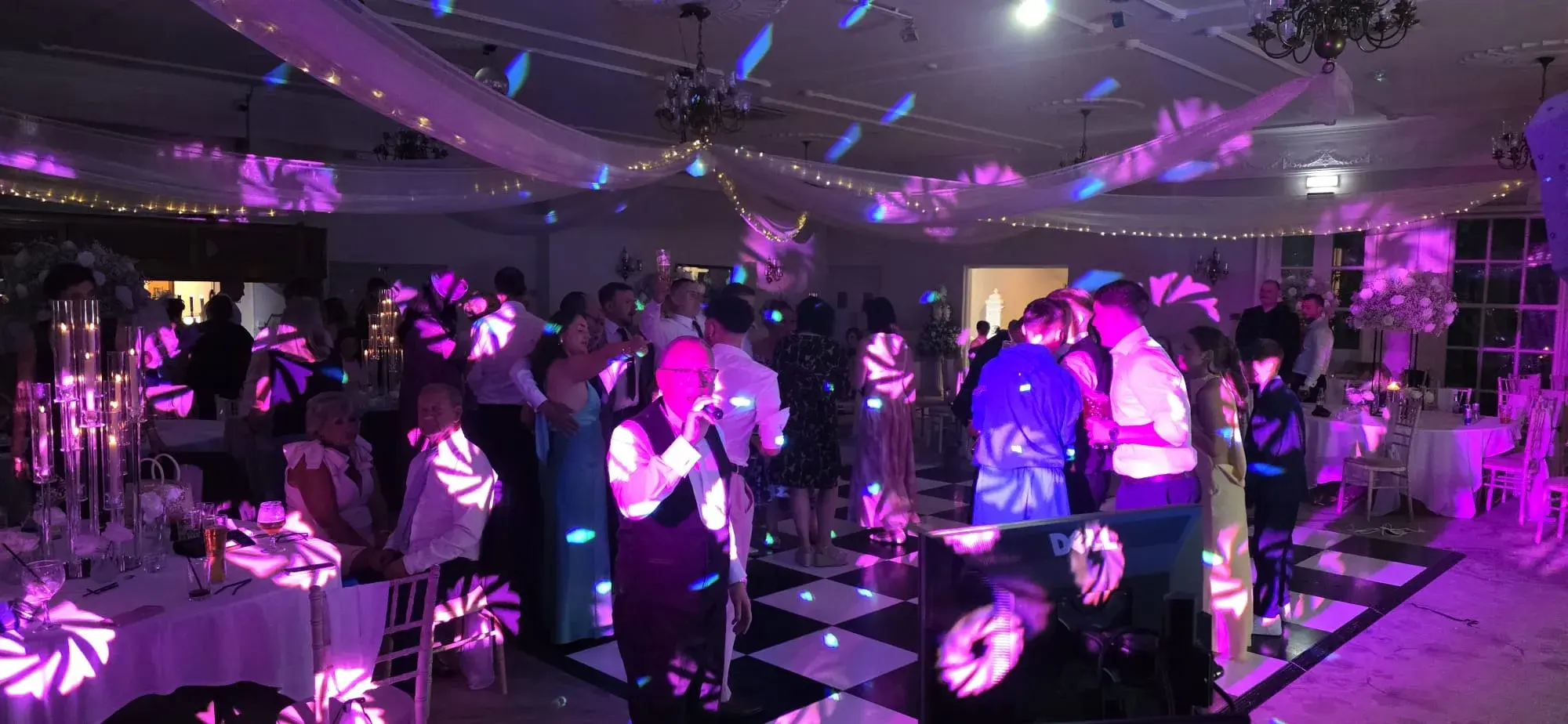 A guest singing into a microphone with wedding guests dancing behind him